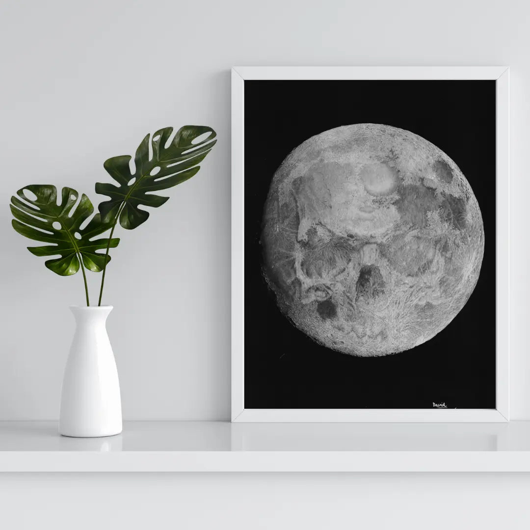 Cuadro enmarcado en blanco de la Luna Calavera, apoyado en una estantería blanca con decoración botánica. Perfecto para decoración gótica, dark o temática espacial.
