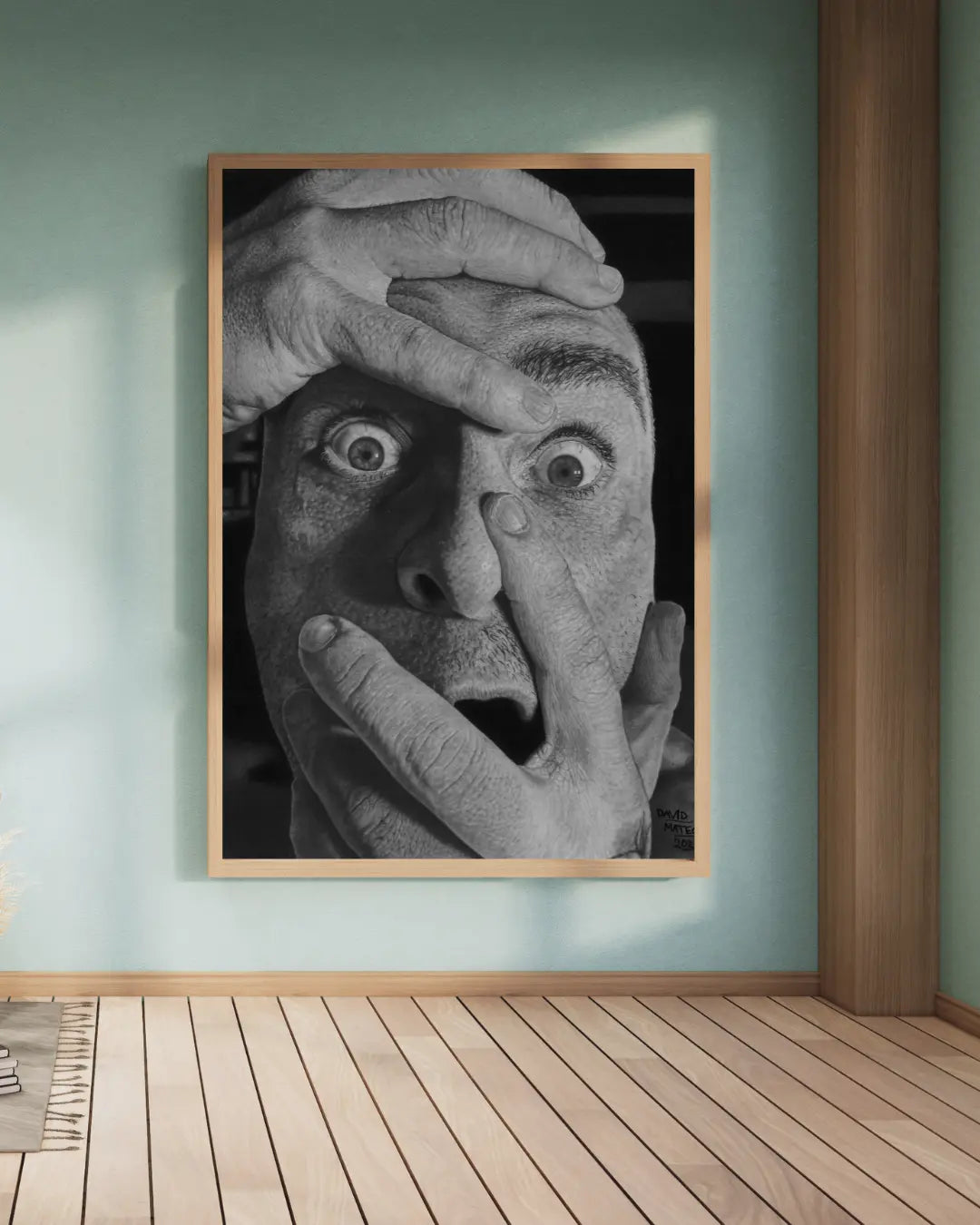 Lámina vertical enmarcada en madera, sobre un escritorio blanco. Muestra la imagen de un hombre en blanco y negro con una expresión de pánico y manos sobre su cabeza y boca.