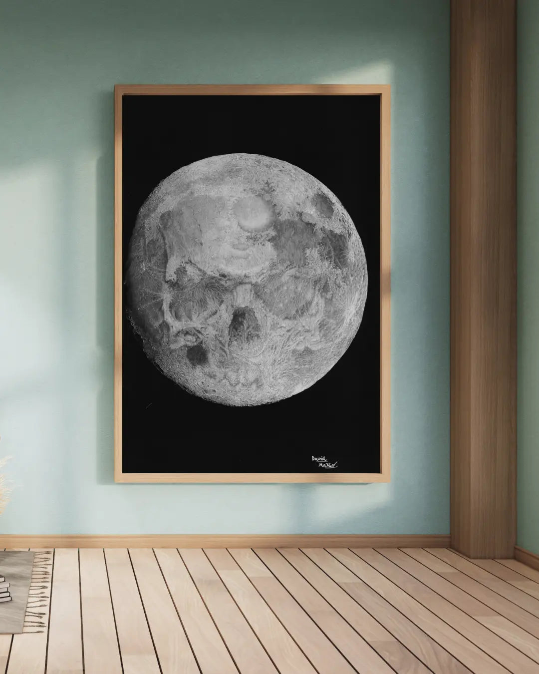 Lámina enmarcada en blanco, sobre una estantería blanca. Muestra un dibujo monocromático de la luna llena en un fondo negro, con un jarrón y una hoja de planta a la izquierda.