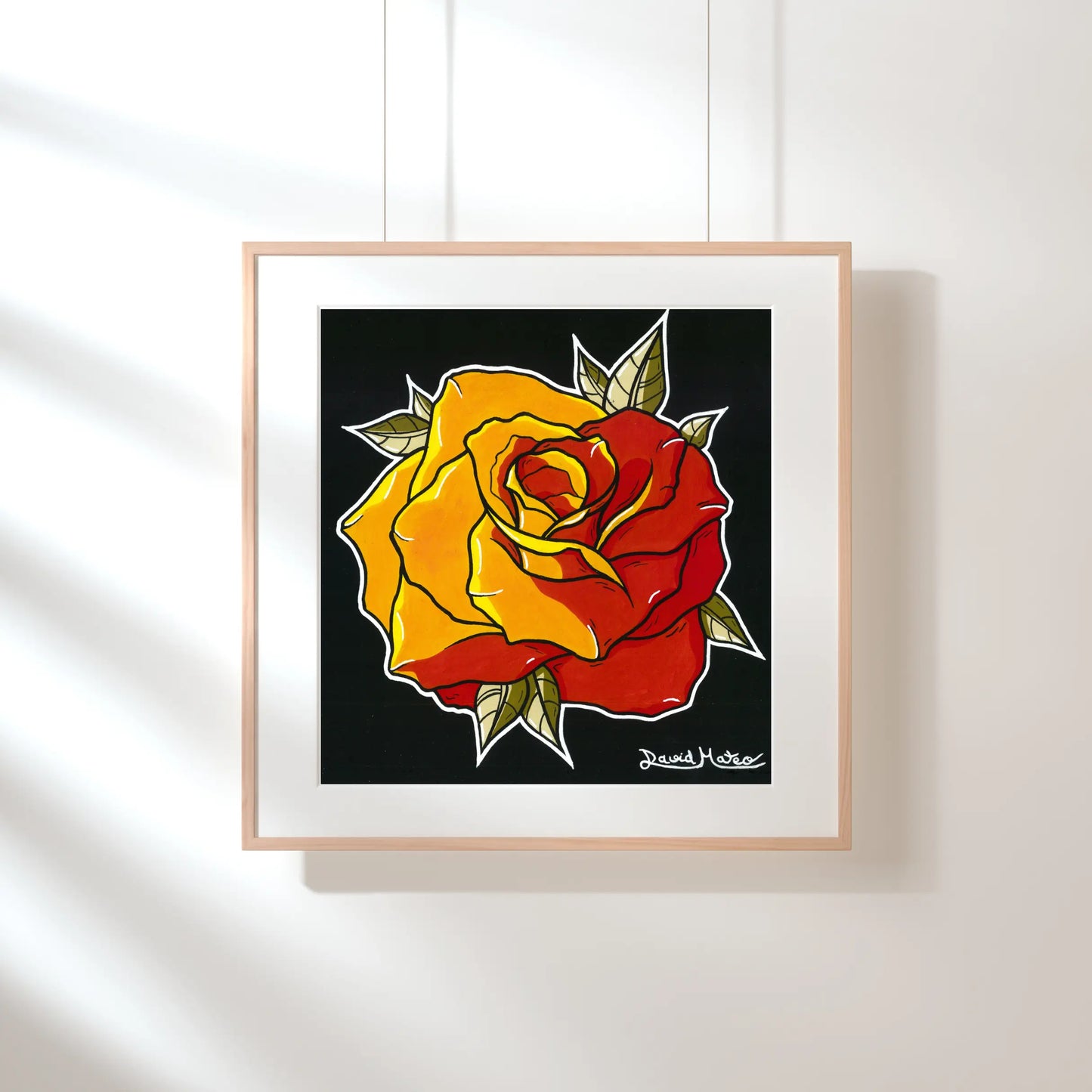 Lámina enmarcada en madera, sobre una pared verde pálido con sombras de luz solar. Muestra el dibujo a color de una rosa de pétalos rojos y amarillos, sobre fondo negro.